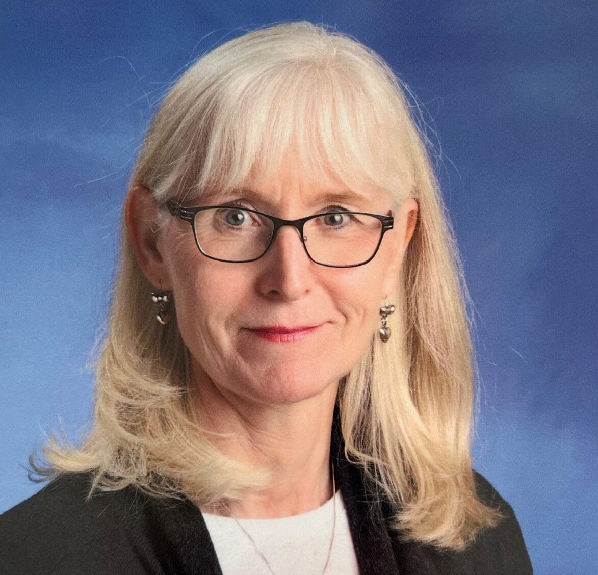 woman with blond shoulder length hair, wearing a whit sweater and black cardigan, she's also wearing rectangular, black framed glasses